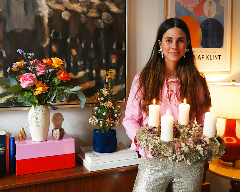 Bloggerin und Influencerin Nike van Dinther hält einen bunten Adventskranz mit brennenden Kerzen in der Hand. Im Hintergrund sieht man weitere farbenfrohen Produkte aus ihrer neuen, für Bloom and Wild kuratierten Geschenke-Edit.