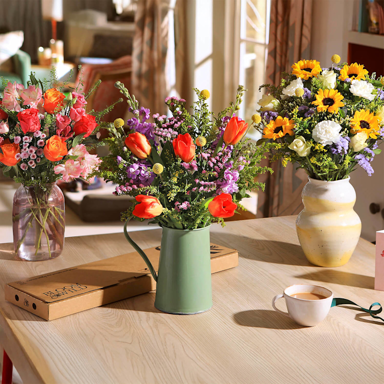 Three vibrant Bloom & Wild bouquets in a sun-drenched room. Features orange tulips in a green jug, bright sunflowers in a ceramic vase, and coral roses in a glass jar. A letterbox flower box and a tea cup sit nearby on a wooden table.