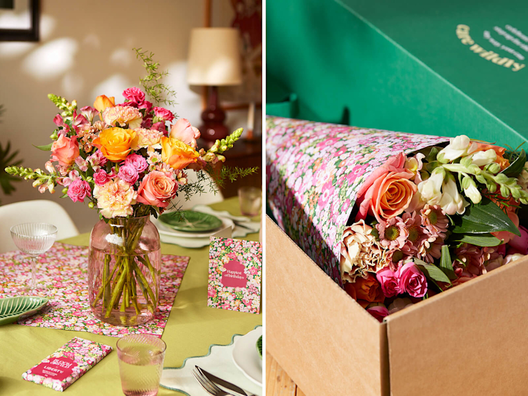 A vibrant bouquet of pink and orange flowers in a clear glass vase sits on a table. The table is covered with a cloth patterned with Liberty print florals, matching a small card and a chocolate bar resting nearby.