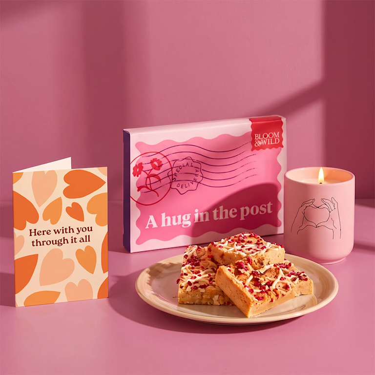 Comforting gift box featuring soft white chocolate raspberry blondie slices, a warm lit pink candle, and a card reading "Here with you through it all".