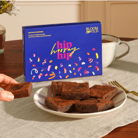 A blue "Hip Hip Hooray" birthday brownie box with colourful confetti design, beside a white plate of six chocolate brownies, with a hand reaching for one and a mug in the background.