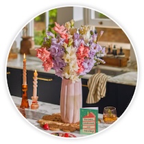 Eine hohe, hellrosa Vase mit einem bunten Strauß aus pastellfarbenen Gladiolen steht auf einer Küchentheke, umgeben von zwei brennenden Kerzen, Gläsern und einer Glückwunschkarte, dargestellt in einem kreisförmigen Bildausschnitt.
