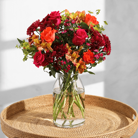 A glass vase of vibrant red, orange, and yellow flowers including roses and freesia, arranged with greenery, standing on a wooden table.
