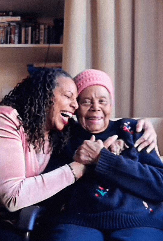 Jaycee, a dark-skinned woman with shoulder-length coiled hair, laughs as she hugs her mum and holds her hands.