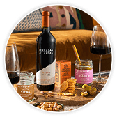 A bottle of red wine, two glasses of red wine, and small jars and packaged snacks arranged on a living room table. 