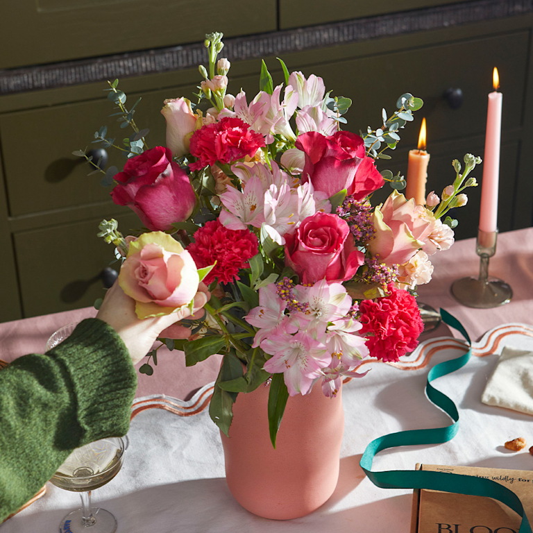A romantic bouquet of deep red and soft pink roses, carnations, and alstroemeria in a matte pink vase. A hand gently adjusts a bloom by glowing candles, evoking a warm, fragrant evening atmosphere with velvety textures.