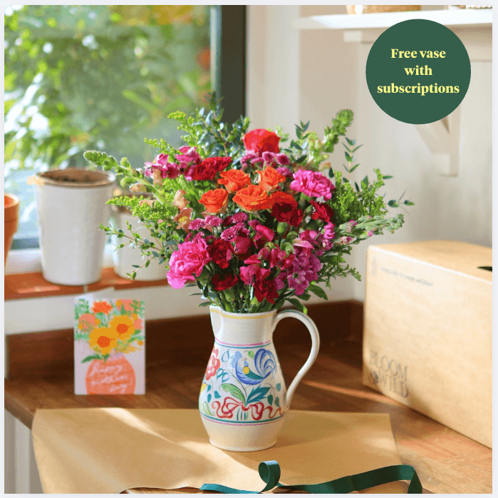 A bouquet of pink roses and white lilies in a clear vase on a round wicker table. A green roundel on the top right of the image reads 'Free vase'.