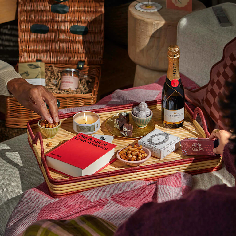 A romantic date night scene featuring a wicker gift hamper, a bottle of Moët champagne, chocolates, and a candle on a woven tray. Two people share a cosy moment, surrounded by the sweet scent of jasmine and treats.