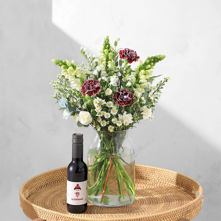 Ein fröhlicher Blumenstrauß mit weißen Blüten und rosaroten Nelken steht in einer Vase auf dem Beistelltisch. Daneben steht eine Piccolo-Flasche mit Glühwein.
