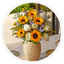 Eine geriffelte, beigefarbene Keramikvase mit einem strahlenden Blumenstrauß aus gelben Sonnenblumen, weißen Rosen und violettem Meerlavendel steht auf einem kariert gedeckten Tisch.