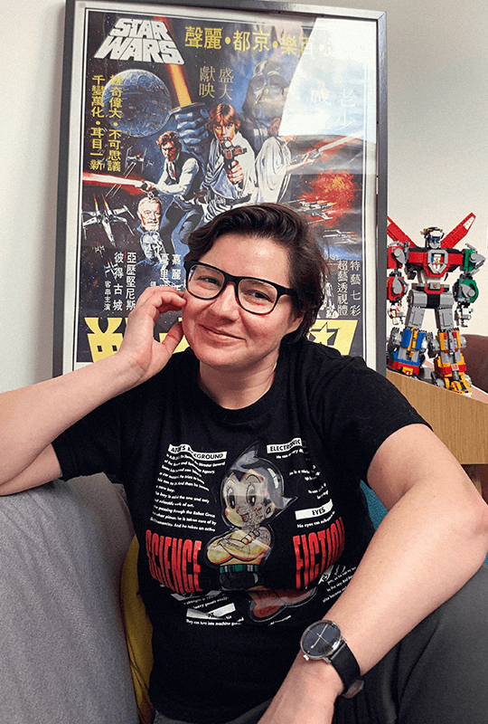 Marta is a light-skinned woman with short-cropped brown hair, wearing a t-shirt and chunky black-framed glasses. She sits in front of an original Star Wars poster. 