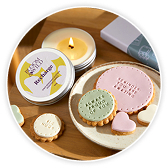 A lit candle in a tin next to round biscuits in pastel pink, green, and cream shades, arranged on a wooden tray.