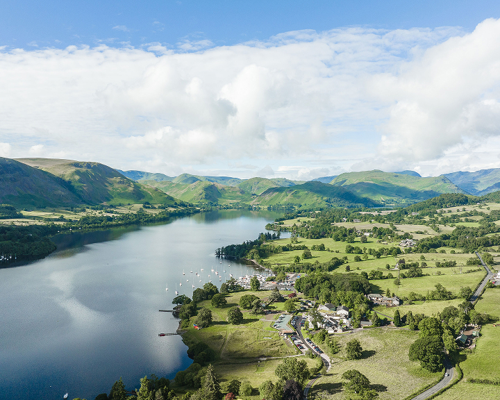 High-angle aerial view of a serene lake in the British countryside under a bright, cloud-dappled sky. Rolling green hills and lush pastures surround the water, dotted with small white sailboats and a winding road.