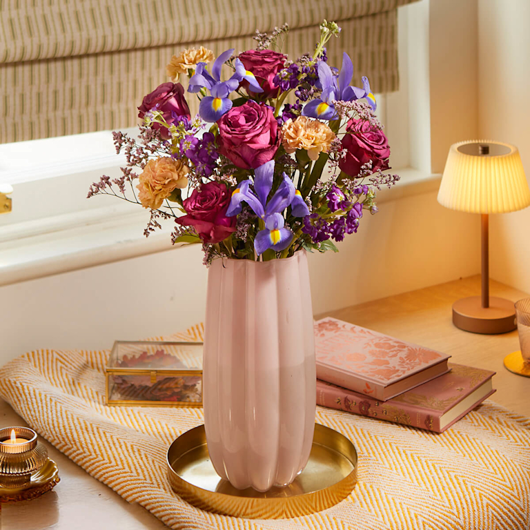 A stunning bouquet of deep purple roses, lilac irises, and peach carnations sits in a rippled pink glass vase. It evokes a feeling of calm and warmth.