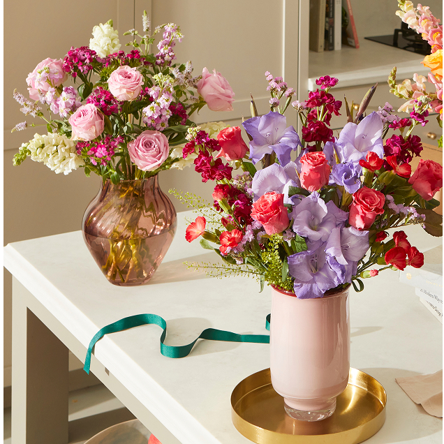 Zwei stilvolle Vasen mit farbenfrohen Blumensträußen stehen auf einem weißen Tisch: Eine hellrosa Vase auf einem Goldtablett mit blau-violetten Gladiolen und korallenroten Rosen, und im Hintergrund eine geriffelte braun-rosa Glasvase mit zartrosa Rosen.