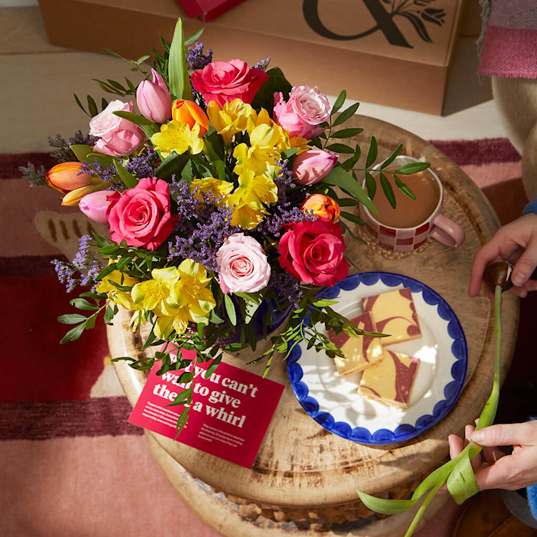 An overhead view of the vibrant Scottie bouquet on a wooden table alongside a plate of marbled brownies and a cup of tea. The bright pink and yellow petals create a cheerful, sweet-scented tea-time treat.