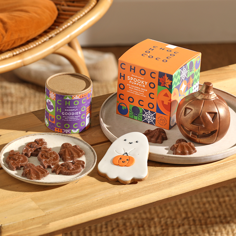 A wooden table holds three Halloween treats. There's a box of 'Spooky Pumpkin' chocolates, a large chocolate pumpkin, and a plate of ghost and bat-shaped chocolates.