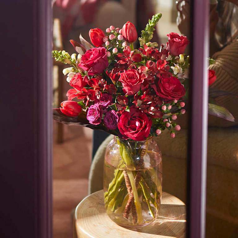 A rich, passionate bouquet of deep red roses, tulips, and pink berries in a glass vase, seen through a doorway. The dramatic crimson tones and lush greenery evoke a sophisticated, fragrant, and romantic feeling.
