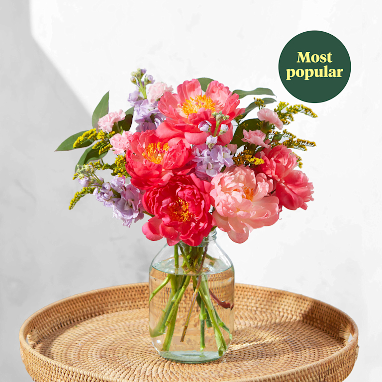 A bouquet of pink and red peonies in a clear vase on a round wicker table. A green roundel on the top right of the image reads 'Most popular'.