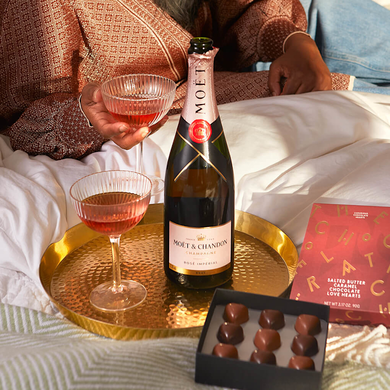 A bottle of Moët & Chandon Rosé Impérial champagne, two glasses of rosé champagne, and a box of chocolates on a gold tray.