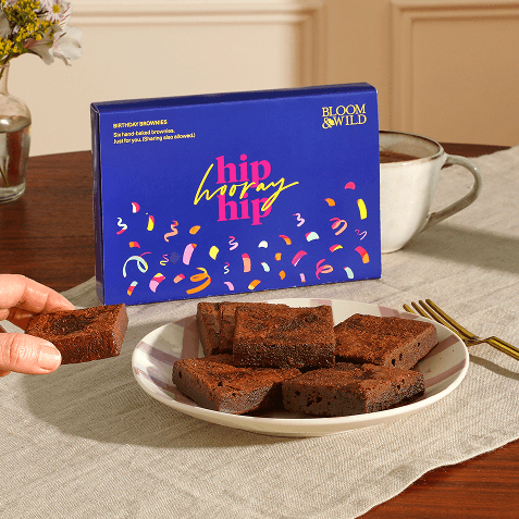 Blue "Hip Hooray Hip" Bloom & Wild brownie box beside a plate of six chocolate brownies, with a hand picking one up, on a dining table.