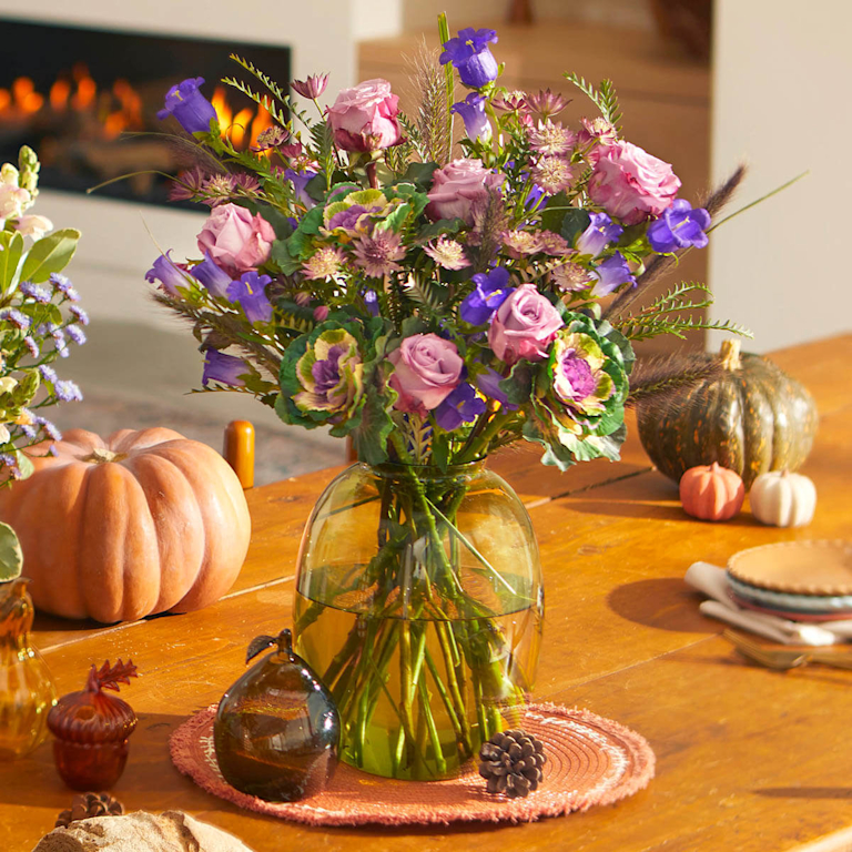 A textured glass vase holds a vibrant bouquet of pink roses and purple bells. It sits on a wooden table, surrounded by pumpkins, with a fireplace in the background.