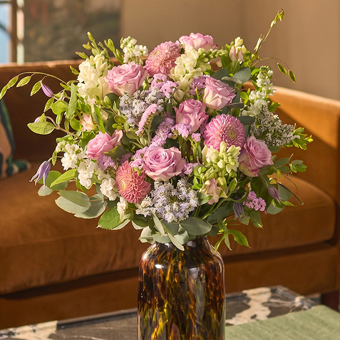 Eine braun gemusterte Glasvase ist mit üppigen Rosen, Dahlien und Löwenmäulchen in Rosa-, Flieder- und Cremetönen gefüllt. Der Strauß steht auf einem Tisch vor einem braunen Sofa in warmem Tageslicht.