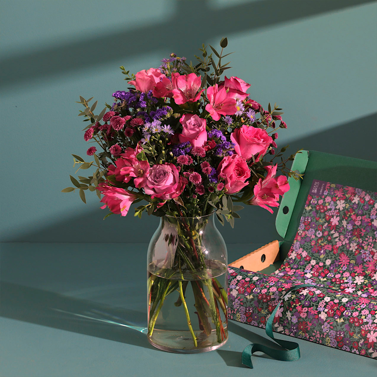 A bouquet of pink roses and purple wildflowers sits in a gray vase on a small table. A woman in a cosy jumper sits in a chair in the background, enjoying a cup of tea.