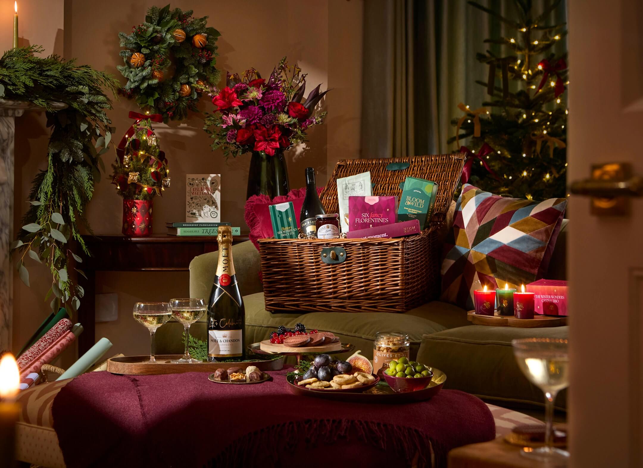 A cosy living room at Christmas with a wicker hamper of festive treats on a green sofa, a bottle of Champagne and glasses, lit red candles, a grazing board on a burgundy footstool and a decorated tree.