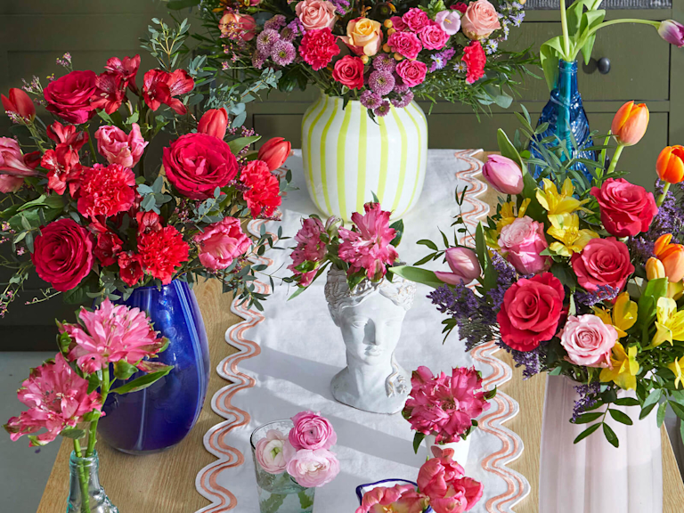 A breathtaking overhead view of multiple Bloom & Wild bouquets and bud vases arranged on a white runner. Vibrant reds, soft pinks, and sunny yellows create a fragrant, meadow-like display that feels abundant and celebratory.