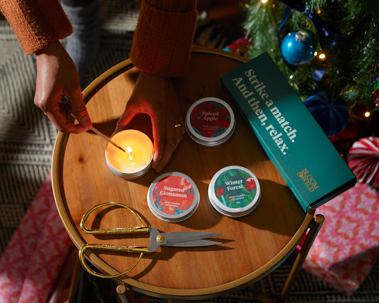 Three Bloom & Wild branded candles in metal tins, being lit by a match. On a table surrounded by a Christmas tree, wrapping paper, and scissors.