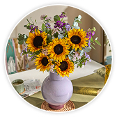 A vase of sunflowers and mixed purple wildflowers arranged in a round white ceramic vase, displayed in a circular frame on a table setting with blurred spring décor in the background.
