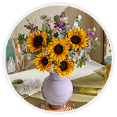 A vase of sunflowers and mixed purple wildflowers arranged in a round white ceramic vase, displayed in a circular frame on a table setting with blurred spring décor in the background.