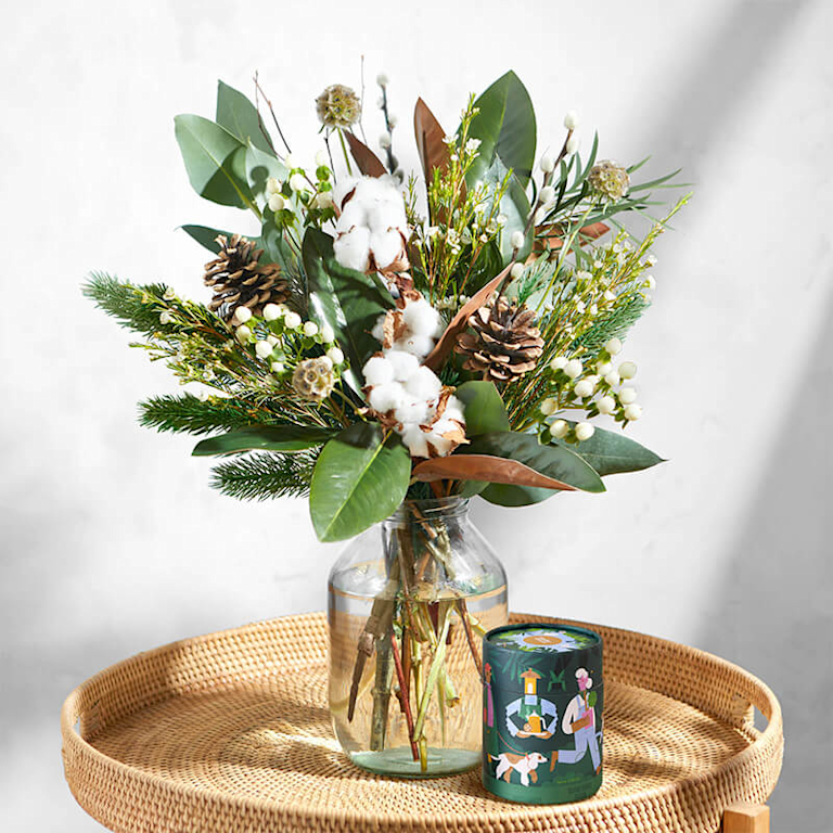 Ein winterlicher, rustikaler Blumenstrauß mit weißen Beeren, grünen Blättern, Tannenzweigen und Tannenzapfen steht in einer Vase auf dem Beistelltisch. Daneben ist eine illustrierte Rundschachtel mit Lebkuchen zu sehen.