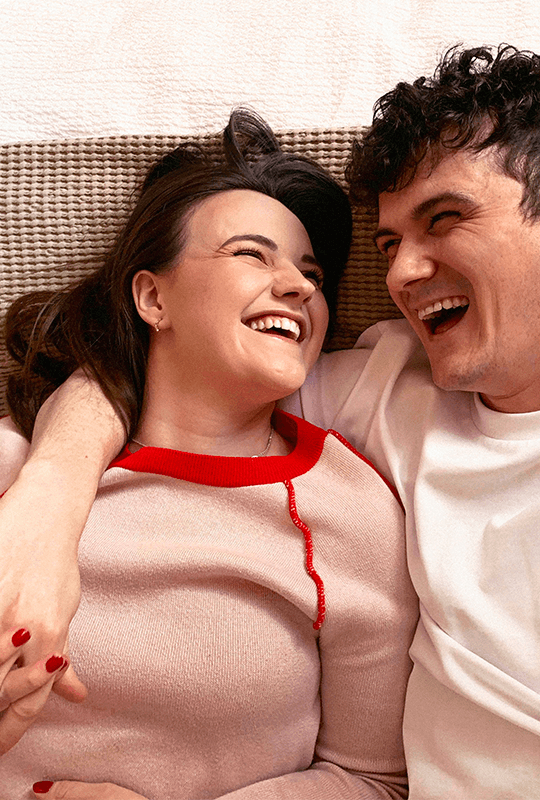 Josh, a light-skinned man with dark curly hair, wraps his arm around his girlfriend Georgia, a light-skinned woman with dark straight hair. Their faces are creased with laughter as they gaze at each other.