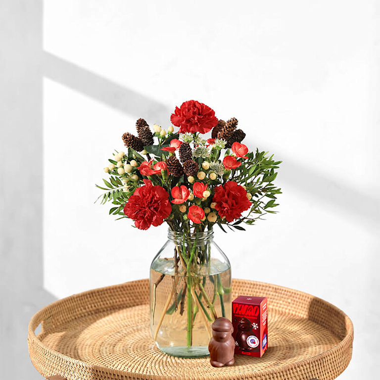 Ein fröhlich festlicher Blumenstrauß mit roten Blüten und Beeren steht in einer Vase auf dem Beistelltisch. Daneben ist ein Schokopinguin von nucao zu sehen.