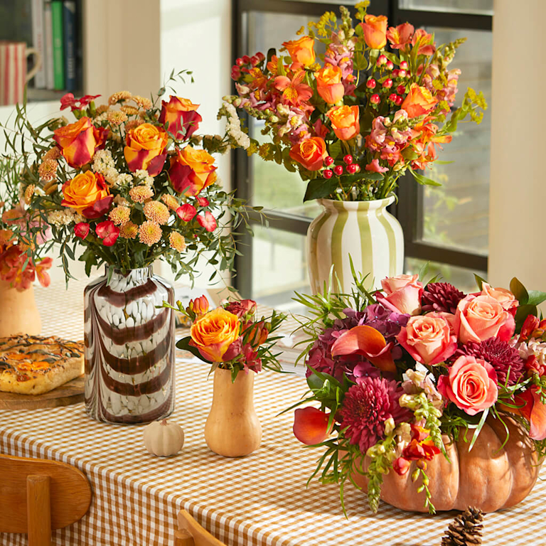 Three autumnal Bloom & Wild bouquets – the Aut and The Orla – on a warmly lit dining table next to a variety of pumpkins and squash in different colours.