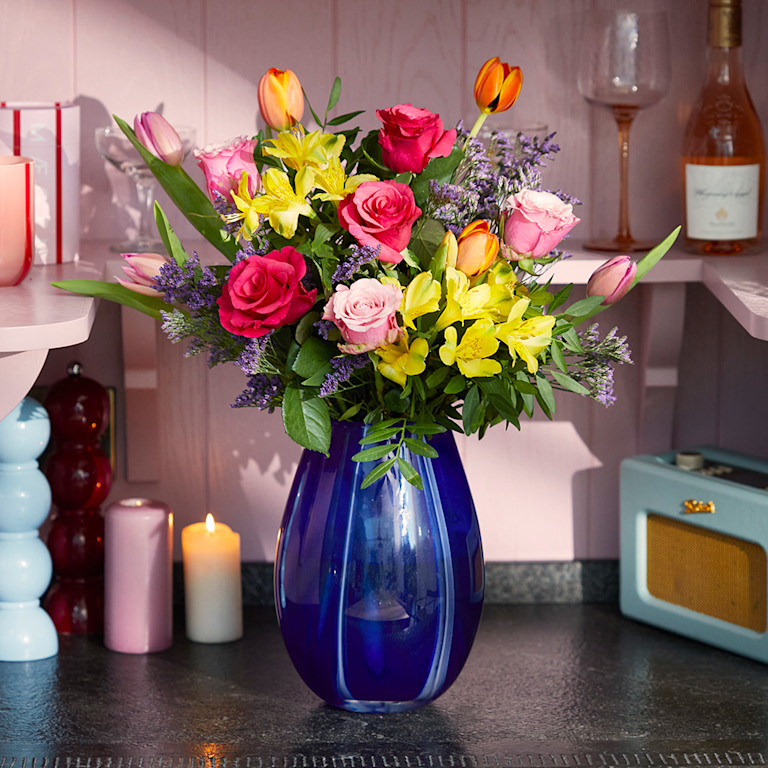 A vibrant, uplifting bouquet featuring hot pink roses, bright yellow alstroemeria, and orange tulips in a striking cobalt blue glass vase. The bold colours and fresh, meadowy scent bring a joyful, sun-drenched feel to the room.