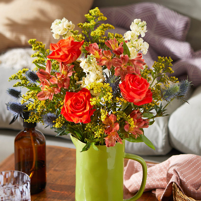 A bright bouquet in a green jug features bold orange roses, alstroemeria, cream stocks, and deep blue thistles. It sits on a wooden coffee table in front of a sofa.