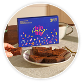 A blue box of brownies labeled "Hip Hip Hooray" beside a stack of chocolate brownies on a white plate, with a hand reaching for one, set on a table with a mug in the background.