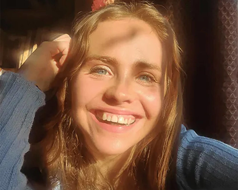 Rosario, a light-skinned woman with wave light brown hair and eyes the colour of sea glass, leans her head on her hand as she smiles widely.