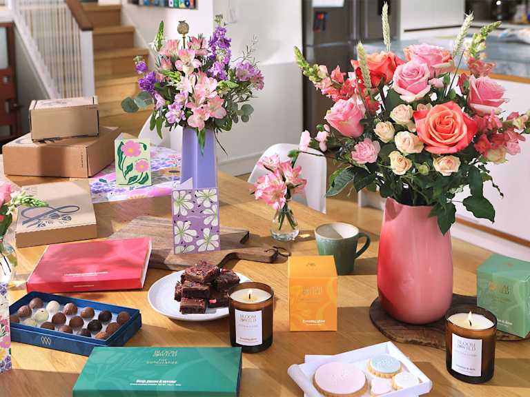 A busy dining table. Two bright bouquets take the main focus. Dotted around them are candles, a stack of mini brownies, iced cookies, chocolate truffles and greetings cards.