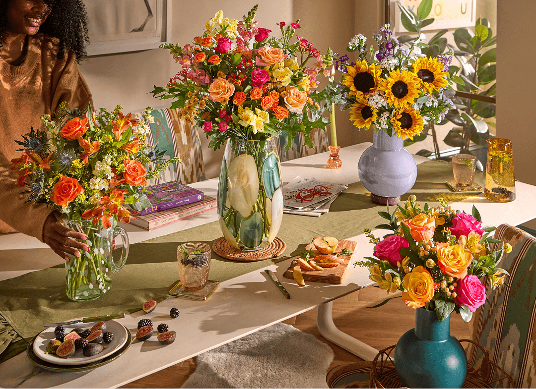 A woman arranges four colorful bouquets—orange roses, mixed pink and yellow blooms, sunflowers, and pink-orange roses—in varied vases on a dining table styled with fruit, books, and a runner in a sunlit room.