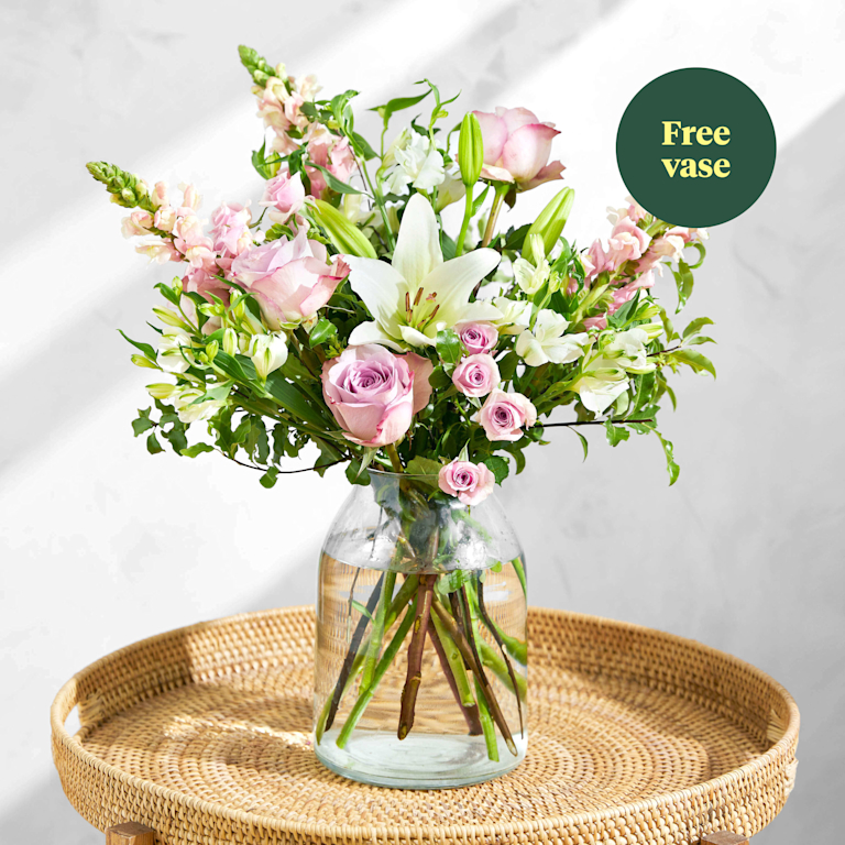A bouquet of pink roses and white lilies in a clear vase on a round wicker table. A green roundel on the top right of the image reads 'Free vase'.