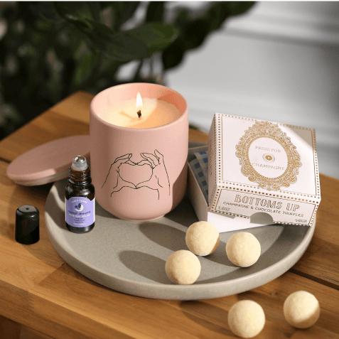Pink scented candle with a heart-hands illustration, a roller-ball fragrance oil, "Bottoms Up" champagne truffles box, and shower steamers on a round tray.