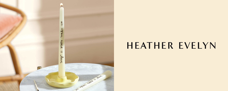 A single tapered candlestick, which is lit, in a scallop shaped ceramic candleholder. A handwritten message on the candle says 'all you need is love'