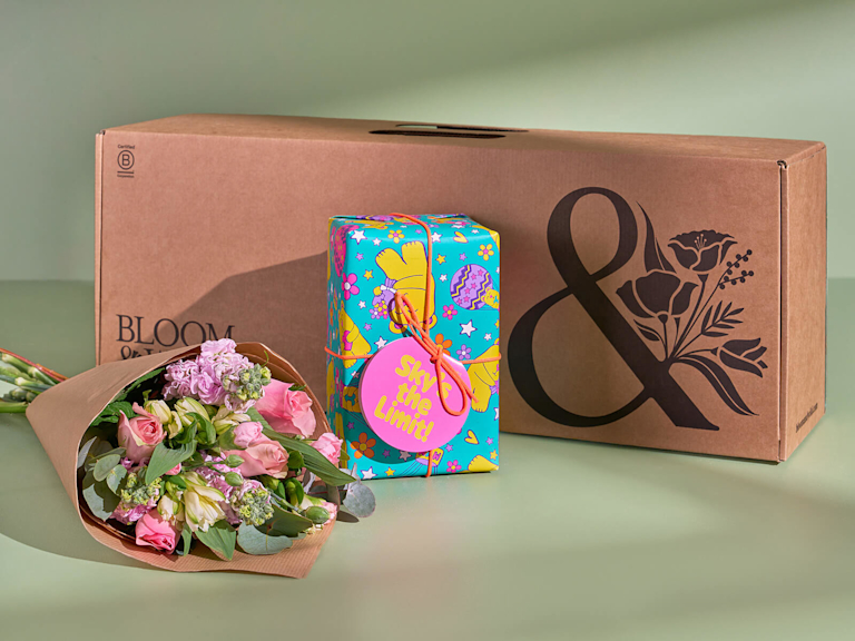 Ein in Blumenpapier eingewickelter, farbenfroher Blumenstrauß. Daneben liegt ein bunt verpacktes Geschenk, indem sich das Geschenkset ‘Sky’s the Limit!’ von LUSH befindet. Im Hintergrund ist die Lieferbox mit dem Bloom and Wild Logo zu sehen. 