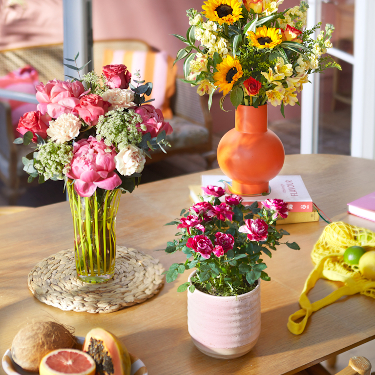 Two bright and colourful bouquets sit on a sunlit kitchen counter. Beside them is premium candle.