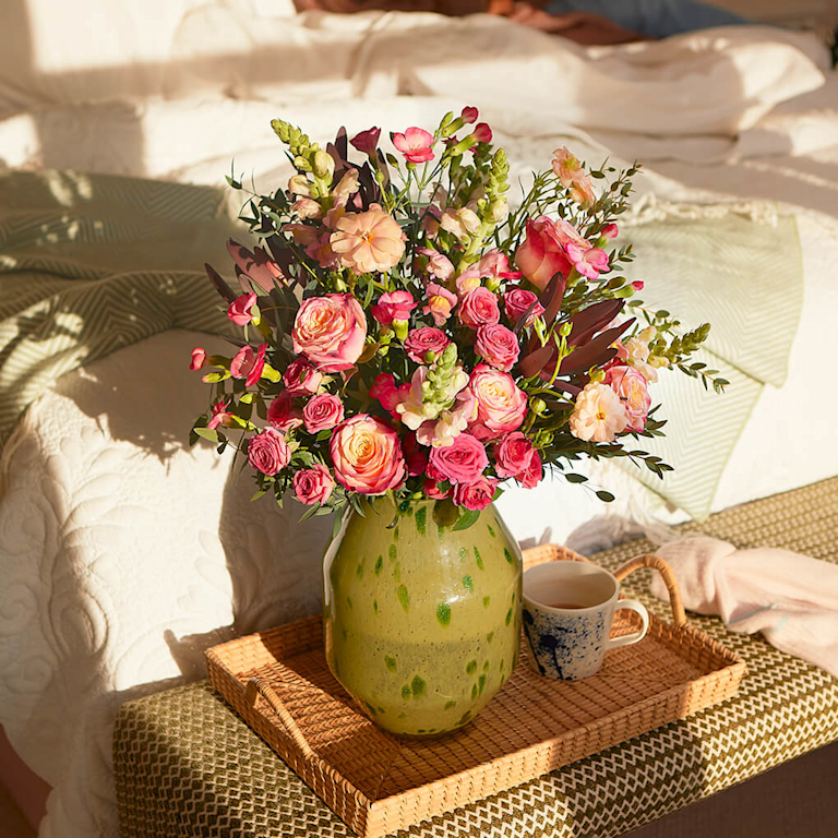 A big bouquet of pink roses, peach spray roses, pink carnations, and pink snapdragons in a green vase on a wicker tray.