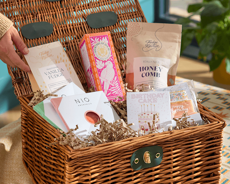 A wicker gift hamper filled with birthday treats including vanilla fudge, shortbread, honeycomb, NIO cocktails, and a "Birthday Cake" gift card, with a hand reaching in and a decorated cake slice visible.